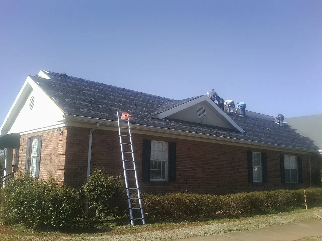 Crew tearing shingles off a brick ranch roof with a ladder set — Emergency Roof Repair work by our crew in Rio