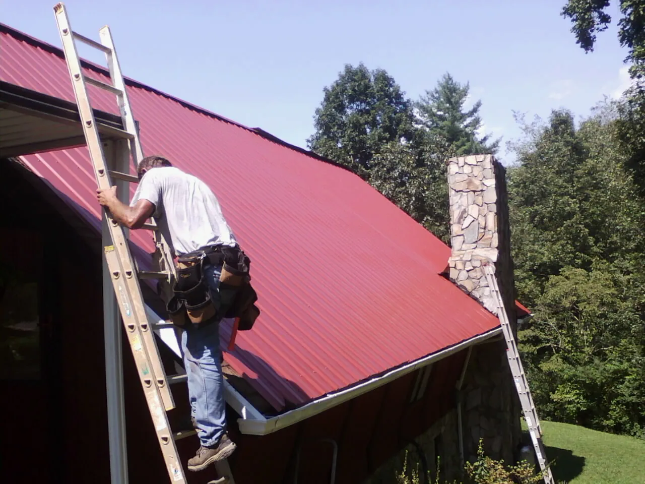 Roofer on a ladder beside a red metal roof and stone chimney — Soffit & Fascia Repair by Roofing Brothers in Rio